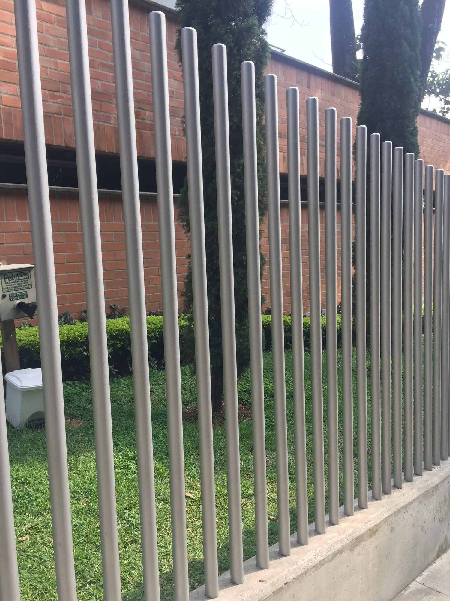 Cerramiento en tubos redondos gris claro frente a un muro de ladrillo